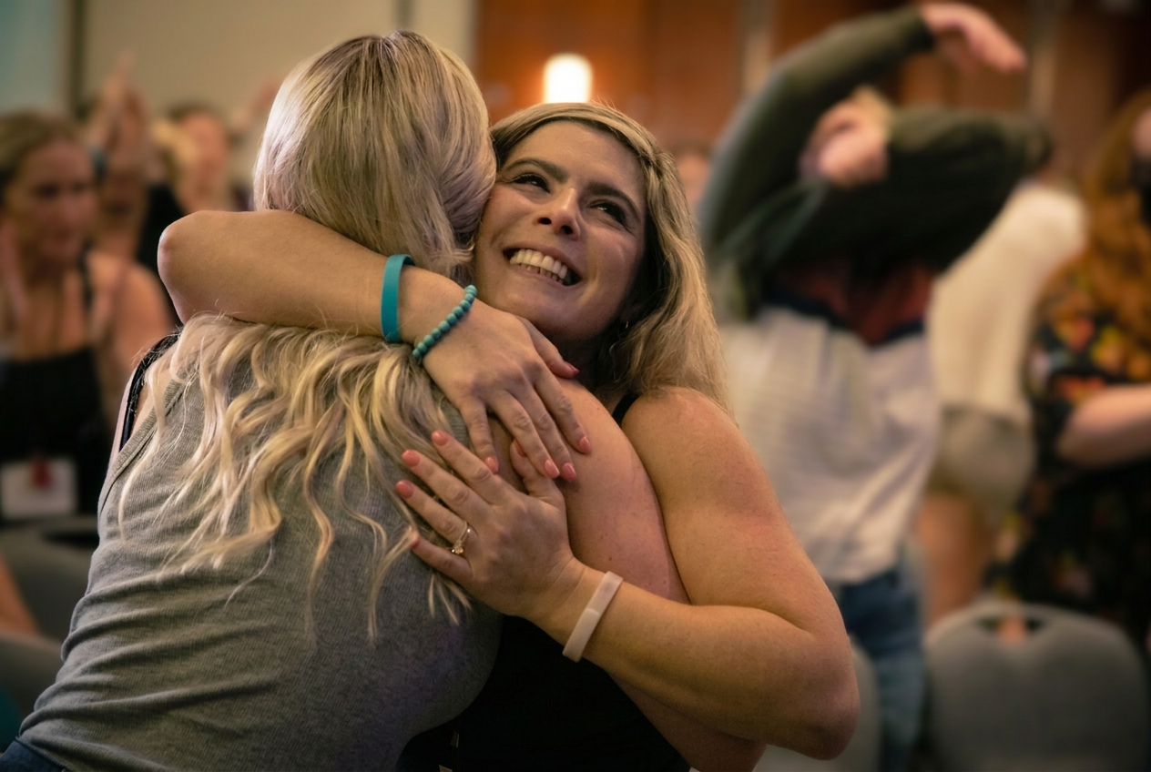 Two event attendees hugging and smiling in a room full of people